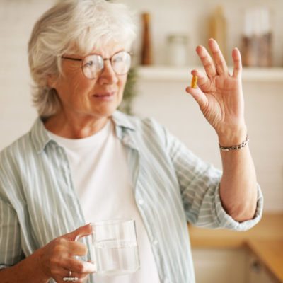 Beautiful Mature Sixty Year Old Female In Stylish Eyeglasses Holding Mug And Omega 3 Supplement Capsule, Going To Take Vitamin After Meal. Senior Gray Haired Woman Taking Fish Oil Pill With Water Beautiful Mature Sixty Year Old Female In Stylish Eyeglasses Holding Mug And Omega 3 Supplement Capsule, Going To Take Vitamin After Meal. Senior Gray Haired Woman Taking Fish Oil Pill With Water