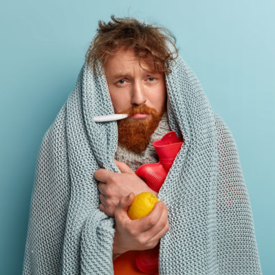 Redhead Young Man With Bristle Fights Against Flu And Fever, Holds Lemon To Enrich Vitamins, Has Thermometer In Mouth, Trembles Under Blanket, Isolated Over Blue Background. Health Problems. Redhead Young Man With Bristle Fights Against Flu And Fever, Holds Lemon To Enrich Vitamins, Has Thermometer In Mouth, Trembles Under Blanket, Isolated Over Blue Background. Health Problems.