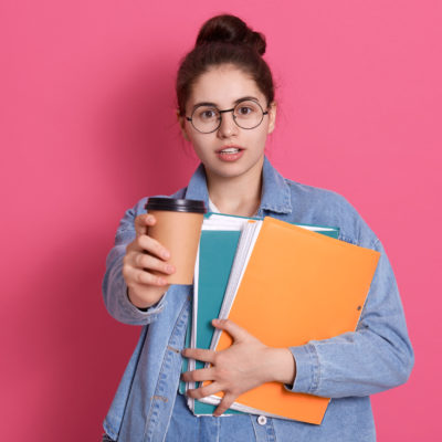Student Girl With Dark Hair Offers Takeaway Coffee, Holding Paper Folder In Hands, Standing Isolated Over Pink Background, Lady Has Coffee Break Between Lectures. Student Girl With Dark Hair Offers Takeaway Coffee, Holding Paper Folder In Hands, Standing Isolated Over Pink Background, Lady Has Coffee Break Between Lectures.